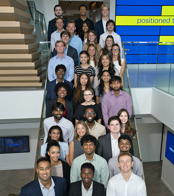 Group of Invesco employees posing together on an office staircase, with a modern interior and digital display in the background.