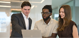 Three Invesco coworkers gather around a laptop, collaborating and smiling in a bright office meeting space.