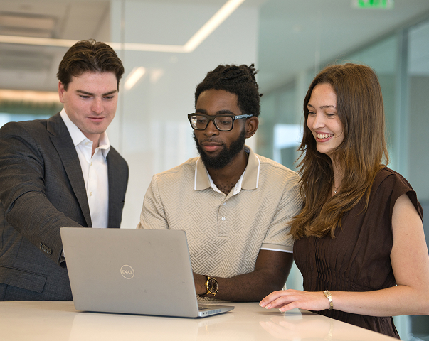 Three Invesco coworkers gather around a laptop, collaborating and smiling in a bright office meeting space.