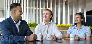 Three Invesco coworkers smile and chat around a table, holding branded mugs in a modern office lounge.