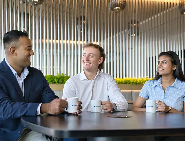 Three Invesco coworkers smile and chat around a table, holding branded mugs in a modern office lounge.